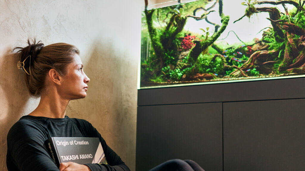 Frau sitzt mit einem Buch vor einem bepflanzten Aquarium und schaut hinein